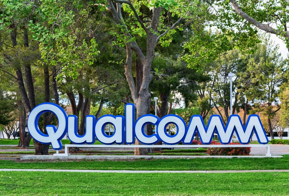 Large "Qualcomm" sign in blue and white letters in front of trees and green lawn.