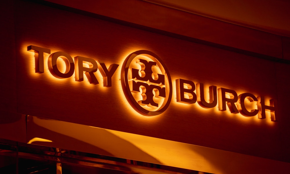 Gold "TORY BURCH" logo with a circular design element on a brown wall with warm lighting.