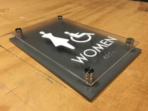 Modern, ADA-compliant office wayfinding sign for a women's restroom, featuring a stylized female figure, a wheelchair symbol, the word "WOMEN", and tactile Braille for accessibility.