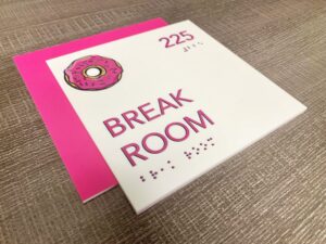 Durable interior sign with a white background and pink accents, indicating "BREAK ROOM" with a donut graphic, room number "225", and tactile Braille for accessibility, suggesting a long-lasting and user-friendly design.