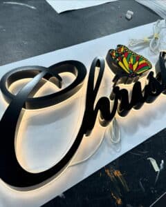 Elegant black dimensional lettering sign in a flowing script with warm white backlighting, featuring a colorful butterfly graphic integrated into the design.
