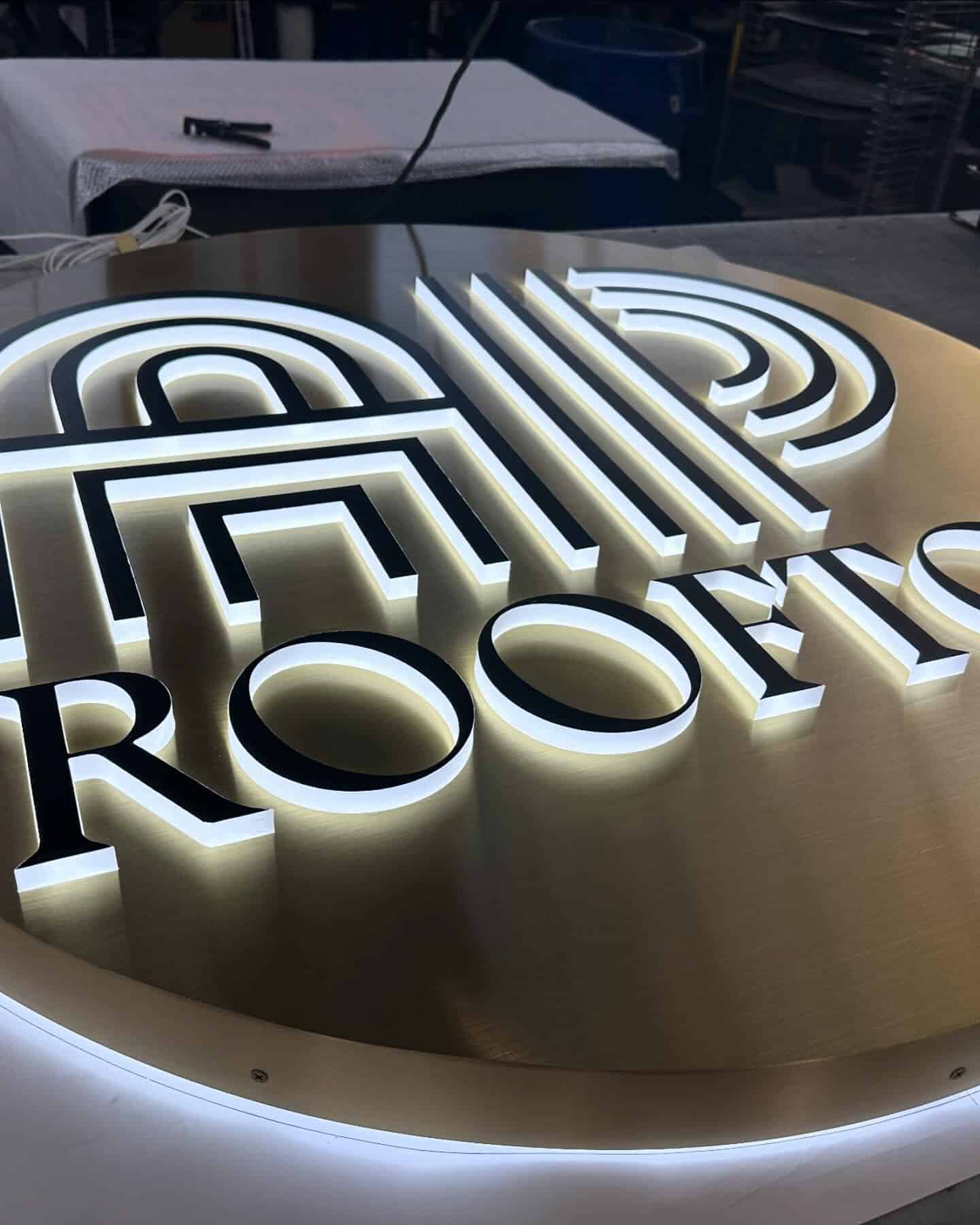 Modern round architectural sign with a gold-toned face, featuring a stylized black and white dimensional logo above the partially visible illuminated lettering "ROOFS"