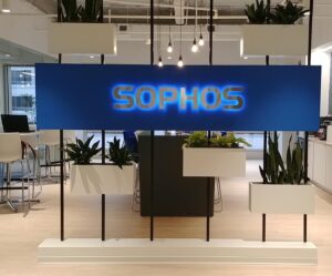 Modern blue corporate lobby sign displaying the word "SOPHOS" in internally illuminated gold dimensional lettering, mounted against a clean backdrop in an office environment with plants.