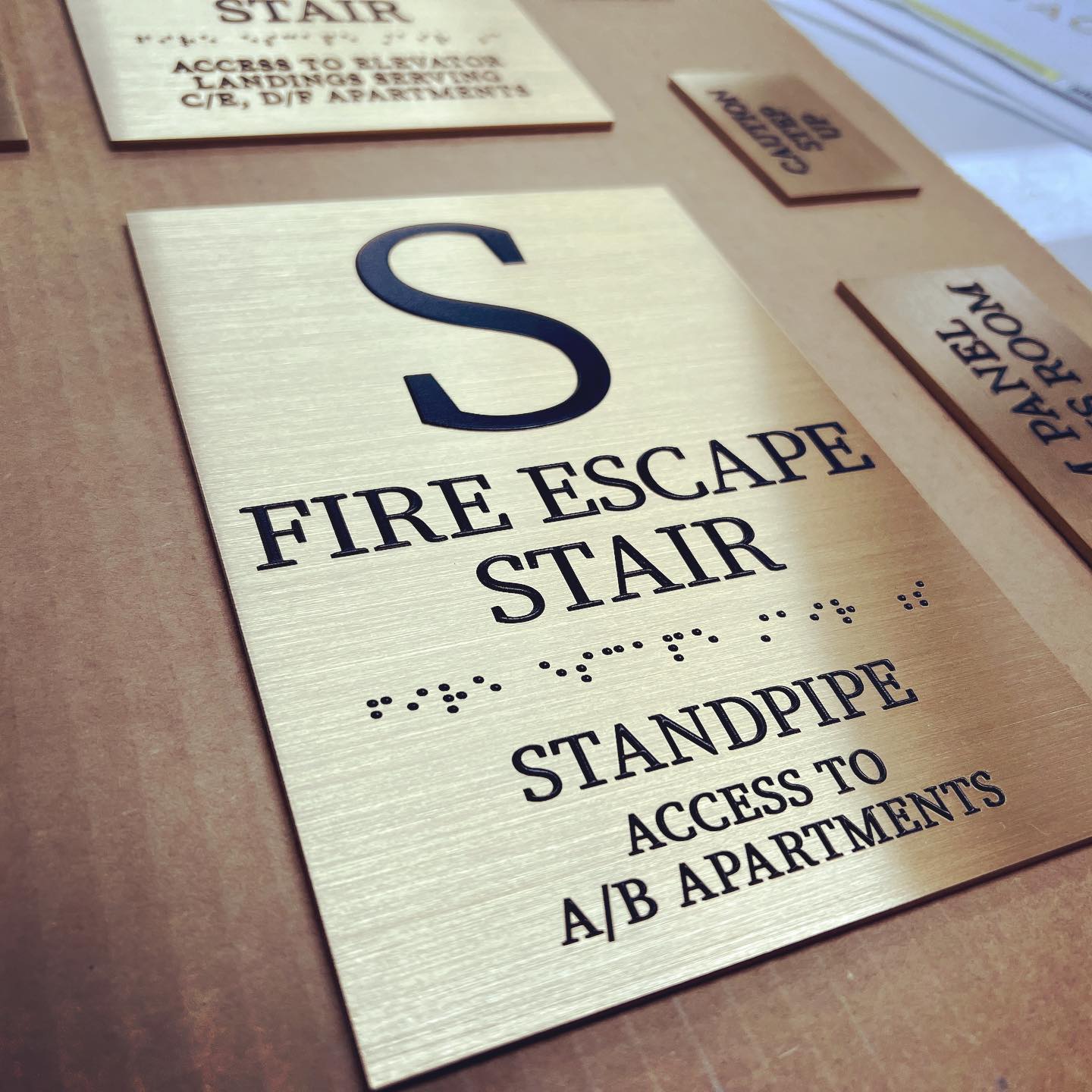 Brushed gold architectural sign indicating "FIRE ESCAPE STAIR" with a large black letter "S" and tactile Braille for accessibility, also noting "STANDPIPE ACCESS TO A/B APARTMENTS.