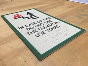 Modern, ADA-compliant office wayfinding sign indicating "IN CASE OF FIRE DO NOT USE THE ELEVATOR. USE STAIRS." with a pictogram and tactile Braille for accessibility.
