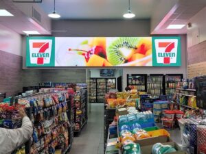 Vibrant LED display above the entrance of a 7-Eleven store, featuring the iconic logo on either side and a colorful image of fresh fruit in the center, illuminating the storefront and attracting attention.