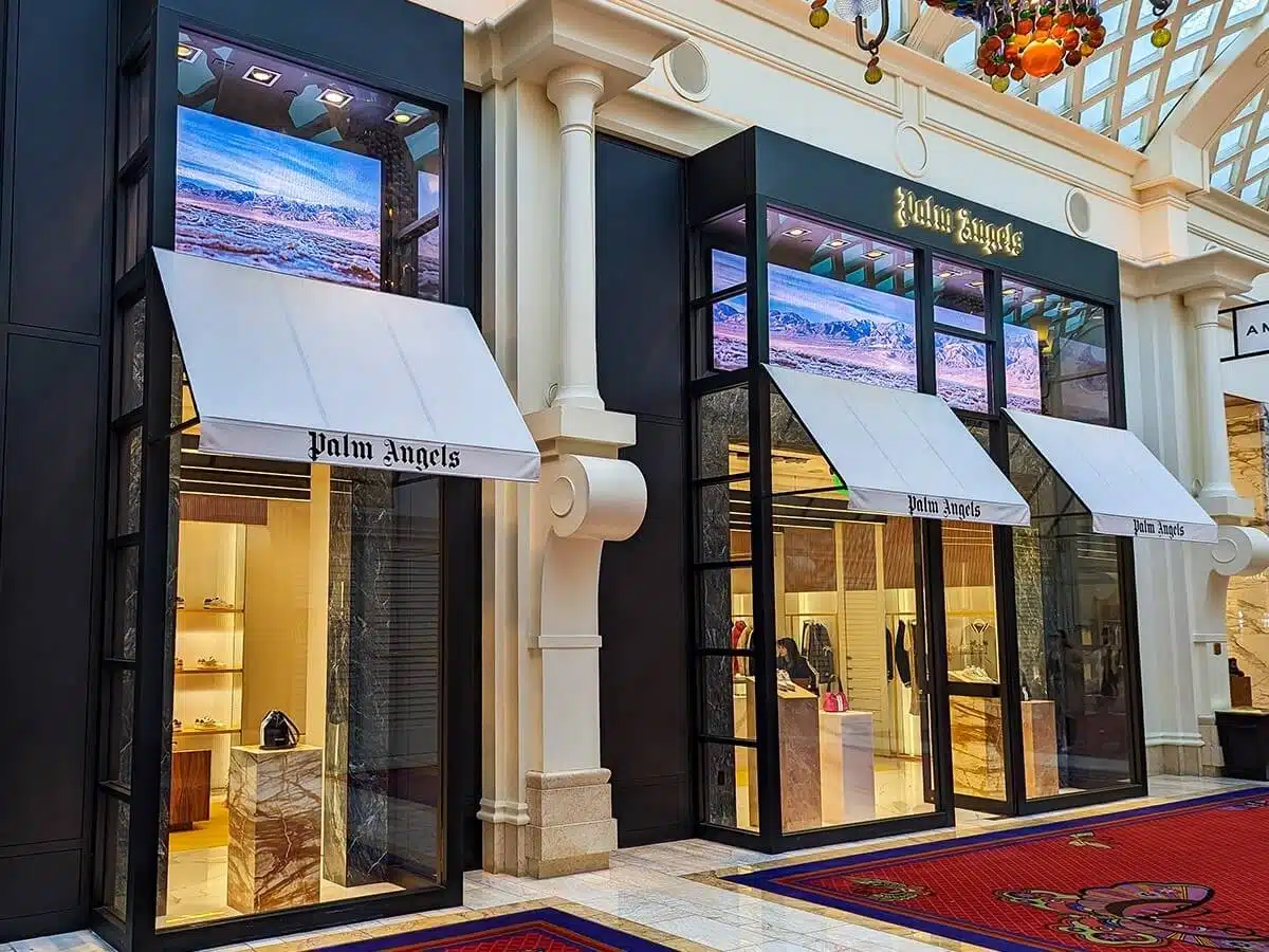 Brightly lit Palm Angels retail storefront with digital displays showcasing scenic mountain visuals above the entrance awnings, enhancing visibility and brand appeal in a shopping area.