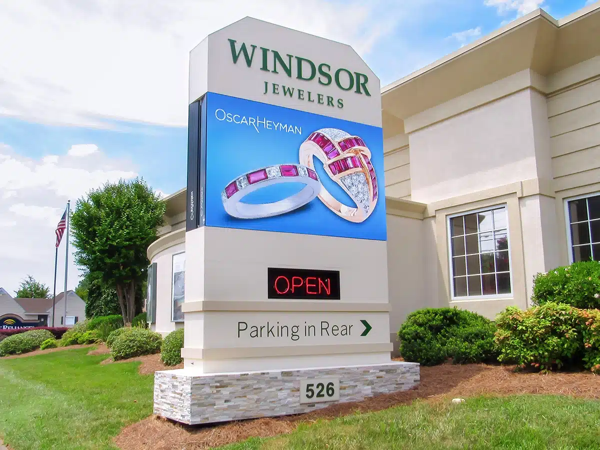Modern monument sign for Windsor Jewelers featuring a digital display showcasing rings, the business name, an "OPEN" sign, and directional information for parking, enhancing visibility and attracting customers to the retail location.